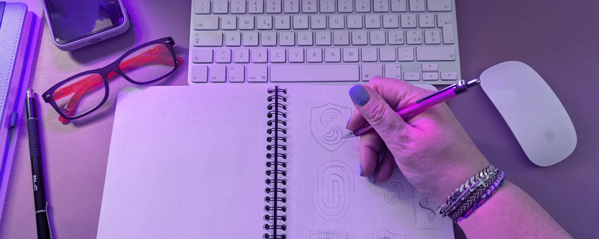 Vista dall'alto della postazione di lavoro di Lisa Creative Mood, brand designer Monza e brianza: computer, tastiera, quaderno lilla e luce LED rosa viola per creare logo e identità visiva.
