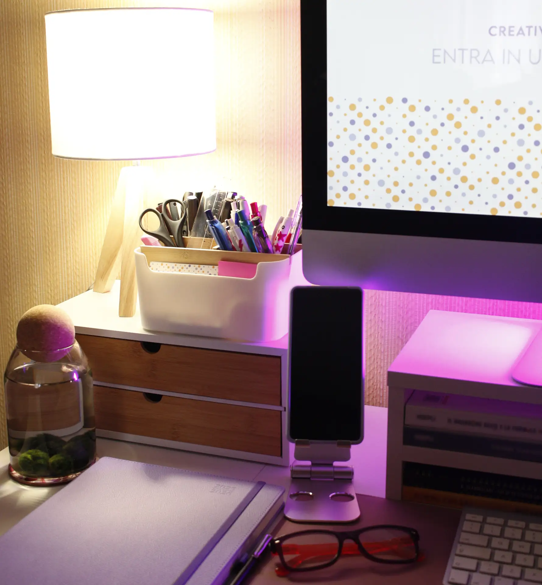 Angolo scrivania con computer, lampada portapenne e cassetti legno sotto luce rosa: allestimento brand designer Monza e brianza per PMI.
