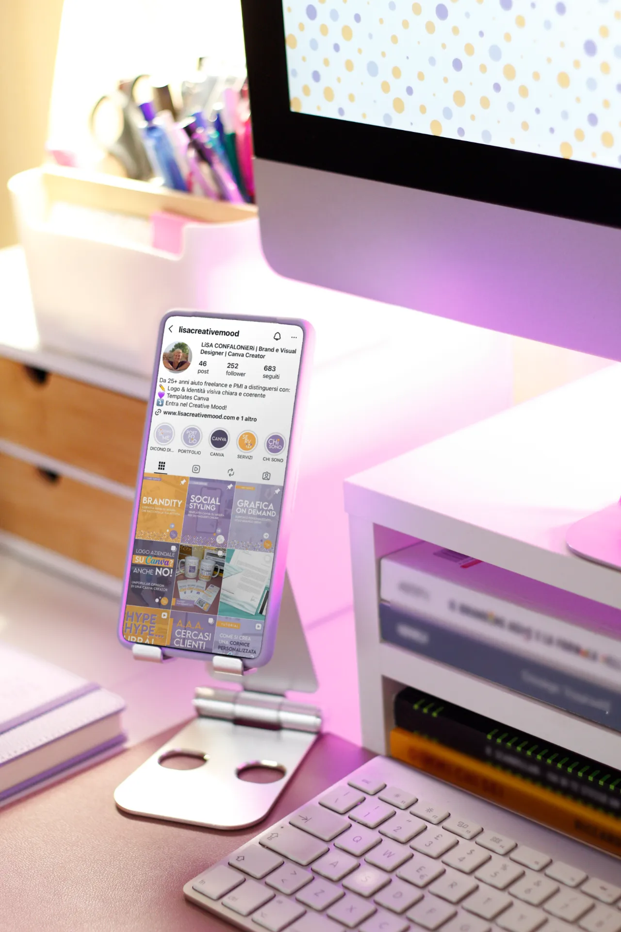 Smartphone su supporto con feed Instagram Lisa Creative Mood davanti a computer su scrivania rosa: ispirazione per brand designer Monza e Brianza e template Canva.